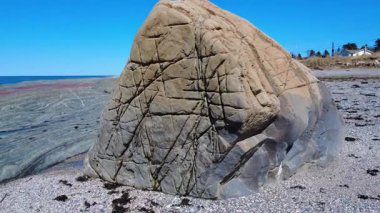 Kanada 'nın Quebec kentinde mavi bir okyanusa uzanan çökelti kayaların önündeki kumlu bir plajda uzanan büyük ve dengesiz bir kayanın etrafında dönen yakın çekim görüntüsü. 2025.