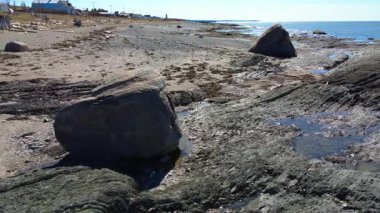 Gözün görebildiği yere kadar uzanan mavi bir denizden yükselen tortulu kayaların yakınındaki kumlu bir plajda duran bir çift dengesiz kaya etrafında dönüyorlar. Baie-des-Sables, Quebec, Kanada, 2025.