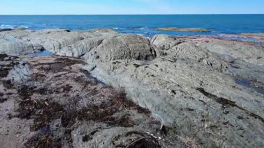 Tortulu bir kaya oluşumu bizi gözün görebildiği kadar uzağa uzanan mavi bir okyanusun dalgalarına götürüyor. Baie-des-Sables, Quebec, Kanada, 2025.