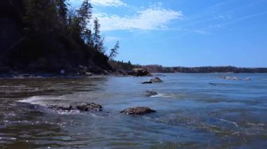 Hızla akan bir nehrin ağzında, ormanın altında, mavi bir gökyüzünün altında sarılmış kayalık kayalıklardan oluşan kıyıya doğru düşük seviyeli bir uçuş. Mitis Nehri, Quebec, Kanada, 2025.