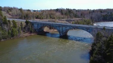 Eski çift kemerli bir yol köprüsünün havadan görünüşü, beyaz bir araba tarafından geçildi, bir somon nehrini bahar ormanından geçirdi. Arthur-Bergeron Köprüsü, Quebec, Kanada, 2025.