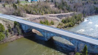 Birkaç arabanın geçtiği, eski çift kemerli yol köprüsünün havadan görünüşü. Bahar ormanlarından geçen bir somon nehri taşıyor. Arthur-Bergeron Köprüsü, Quebec, Kanada, 2025.