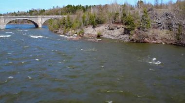 Bir kampçı, ormanla çevrili aşınmış, güçlü bir nehri taşıyan çift kemerli beton bir köprüyü geçer. Arthur Bergeron Köprüsü, Quebec, Kanada, 2025.