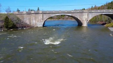 Bir kaya, eski bir beton köprünün kemerinin altından akan bir nehrin yüzeyinde girdaplar yaratır. Mitis Nehri, Arthur-Bergeron Köprüsü, Quebec, Kanada, 2025.