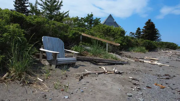 Bereketli bir kumsaldaki ahşap bir sandalyenin sakin sahnesi yemyeşil ve ağaç dallarıyla çevrili bir ev çatısının koçanı bitkilerden dışarı bakıyor ve temiz bir gökyüzünü işaret ediyor..