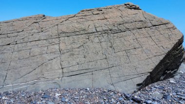 Yıpranmış jeolojik bir kaya oluşumunun düz bir yüzü görünür çatlaklarla ona mavi gökyüzünün altındaki çakıl taşı plajında antik bir taş görünümünü verir..