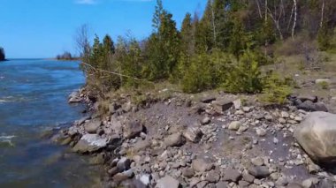 İHA videosu, bulutsuz mavi bir gökyüzünün altında, ormanlık bir nehrin kayalık kıyısından uzak okyanusa uçuyor. Mitis Nehri, Quebec, Kanada, 2025.