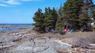 Üç kırmızı ve mavi sandalye ağaçların altında küçük bir ormanlık alanda dizilmiş ve mavi bir gökyüzünün altında, St. Lawrence Nehri 'nin dalgalarla parçalanmış kayalık kıyılarını inceliyorlar. Mtis-sur-Mer, Quebec, Kanada, 2025.