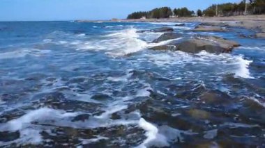 Düşük seviyeli insansız hava aracı dalgaların üzerinden sahilin yakınındaki kayalık bir sahile çarpıyor. Arka planda bir orman ve mavi gökyüzü var. Metis-sur-Mer, Quebec, Kanada, 2025.