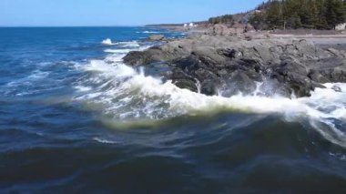 Dalgaların kıyıya vurduğu kayalık kıyı manzarası açık mavi bir gökyüzü ve arka planda yemyeşil bir orman. Vahşi güzelliğin ve dinamik kıyı manzarasının büyüleyici bir tasviri. Metis-sur-Mer, Quebec, Kanada, 2025.