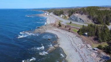 St. Lawrence Nehri sahil şeridi panoramik hava manzaralı kayalık bir plaj, ormanlık bir alan ve açık mavi bir gökyüzünün altında kıyı boyunca uzanan dolambaçlı bir yol ufuktaki uzak bir rüzgar çiftliğine. Quebec, Kanada, 2025.