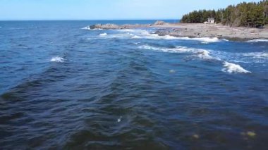 Küçük, ıssız bir kulübe, mavi bir gökyüzünün altında kayalık bir sahile çarpan ve köpüren dalgalarla karşı karşıya. Mtis-sur-Mer, Quebec, Kanada, 2025