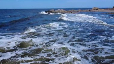 Bir dron, mavi gökyüzünün altındaki kayalık bir noktaya çarpan ve köpüren bir dizi dalgayı gözün görebildiği yere kadar uzanan bir okyanusu takip eder. Mtis-sur-Mer, Quebec, Kanada, 2025.