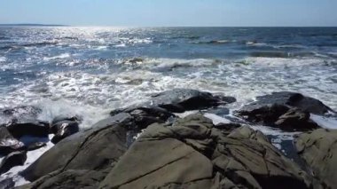 Açık mavi bir gökyüzünün altında okyanus dalgalarının sıçradığı kayalık bir sahil manzarası. Güneş ışığı sudan yansıyarak rahatlatıcı ve dingin bir ortam ekler. Metis-sur-Mer, Quebec, Kanada.