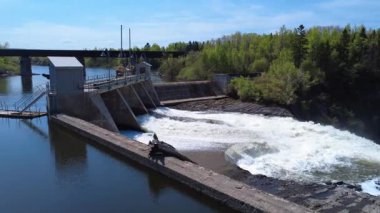 Arka planda demiryolu köprüsü olan ormanın kenarındaki Mitis-1 hidroelektrik santrali barajının vanalarının ve taşkın duvarlarının üzerinden insansız hava aracı uçuşu. Price, Quebec, Kanada.