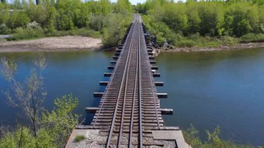 Nehirden geçen bir demiryolu köprüsünün yüksek görüntüsü, her iki tarafında canlı yeşillik ve sudaki sakin yansımalar, doğada bağlantı ve manzaralı seyahat fırsatlarını temsil ediyor..