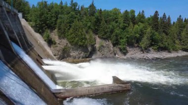 Canlı yeşil bir orman ve dik kaya oluşumlarıyla çevrili barajdan su salan bir hidroelektrik santralinin statik videosu yenilenebilir enerji sergiliyor. Mitis-2 Elektrik Santrali, Fiyat, Quebec, Kanada, 2025.