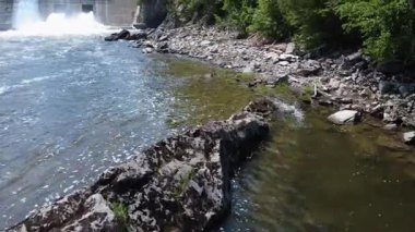 Ormanla çevrili bir nehir ve kayalık kıyısı üzerinden hidroelektrik santrali baraj duvarının su boşaltım kanallarına doğru uçuyor. Mitis-2, Price, Quebec, Kanada.