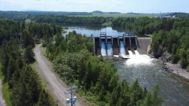 Artan su ve tam akış kapaklı bir hidroelektrik baraj manzarası, endüstriyel tasarım ve bereketli bir orman ortamının birleşimi, sürdürülebilir yenilenebilir enerji üretimi sergiliyor. Mitis-2 Elektrik Santrali, Fiyat, Quebec, Kanada.