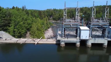 Berrak mavi gökyüzü altındaki yoğun yeşil ormanların ortasındaki nehir seviyesini ve akışını kontrol eden modern baraj kapısı yapısı, sürdürülebilir su yönetimi ve doğanın güzelliğini vurguluyor. Mitis-2 Barajı, Quebec, Kanada.