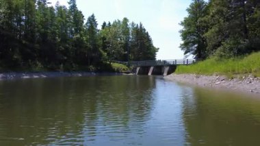 Köprünün altından geçen iki beton kirişin arasından geçen bir dron tarafından çekilen video beton duvarla çevrili bir göle ve mavi gökyüzünün altındaki ormanın üzerine çıkıyor..