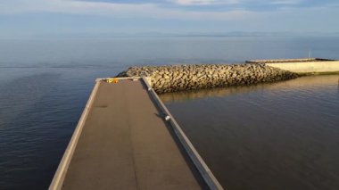 Huzurlu bir manzara, sakin bir okyanusa uzanan kayalık, beton bir iskele, dinlenmek ve güneşin sakin atmosferinin tadını çıkarmak için huzurlu bir yer sağlayan iki renkli sandalye..