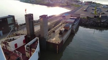 İskele altyapısı, sabah güneşinin altında sakin sularda yüzen geminin pruvasına giden beton bir giriş koridorundan araçları almaya hazır bir feribotla kenetlendi. Riviere-du-Loup, Quebec, Kanada, 2025.