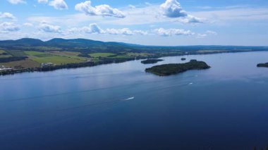 Bir grup zevk botunun panoramik hava görüntüsü bulutlu bir gökyüzünün altındaki dağlık bir tarım vadisi boyunca uzanan geniş bir tatlı su gölünün yakınındaki bir adaya doğru gidiyor. Matapedia Gölü, Quebec, Kanada.