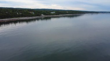 Sahil, evler, ağaçlık alanlar ve bulutlu bir gökyüzünün altında sakin suların bulunduğu sakin bir sahil şeridinin panoramik klibi. St. Lawrence Nehri, Quebec, Kanada.