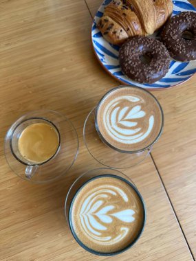 Lezzetli kahve ve tatlı seçeneklerle süslenmiş güzel bir masa kompozisyonu. Görüntü, bir espresso ve bir tabak kruvasan ve donutla birlikte iki kapuçino da dahil olmak üzere üç tip kahve yakalıyor. Sıcak ve davetkar.