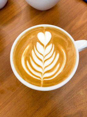 Üzerinde güzel bir süt sanat tasarımı olan bir fincan latte, üzerinde kalpler ve yapraklar, ahşap bir masanın üzerinde dinleniyor..