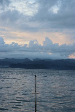 Banliyölerde sakin bir sahil manzarası, sakin deniz suyu, bulutlar, dağlar, ağaçlar ve öğleden sonra güzel gökyüzü.