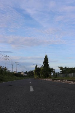 Yol kenarındaki yeşil ağaçların ve yaprakların gölgeli bulutların ve mavi gökyüzünün görüntüsü.