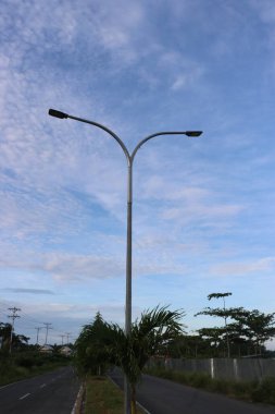 Yol kenarındaki yeşil ağaçların ve yaprakların gölgeli bulutların ve mavi gökyüzünün görüntüsü.