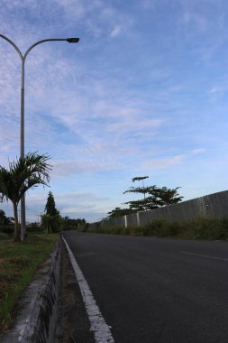 Yol kenarındaki yeşil ağaçların ve yaprakların gölgeli bulutların ve mavi gökyüzünün görüntüsü.