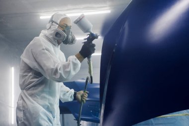 Erkek endüstriyel işçiler, arabanın koruyucu maske ve atölye garajında giyilen kıyafetlerin bir kısmına hava fırçası tabancası kullanarak boya sıkarlar. Adam kaputa boya püskürtüyor.. 