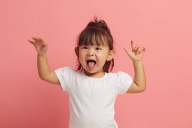 Stüdyo portresi, neşeli küçük Asyalı kızın dilini göstermesi, ellerini kaldırarak ağzını açması pembe izole bir arka planda durması.. 