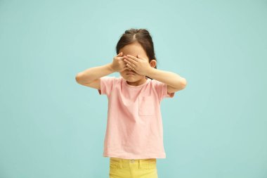 Stüdyo, pembe tişörtlü tatlı anaokulu kızını elleriyle gözlerini kapatıp saklambaç oynarken çekti. Peekaboo 'da oynayan Asyalı bir çocuğun izole edilmiş portresi mavi arka planda duruyor.