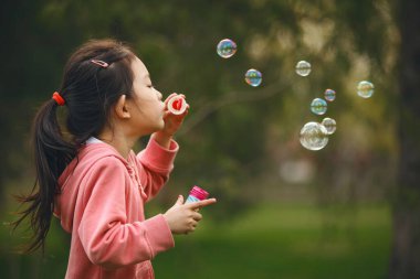 5-6 yaşlarında, koyu saçlı, pembe kazaklı, elinde değnekle köpükler saçan bir kız. Arkasındaki havada birden fazla baloncuk uçuşuyor, ve arka plan bulanık, yeşil