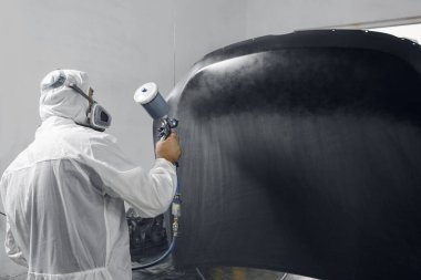 Koruyucu eldivenli sprey boyacısı, boya odasındaki araba gövdesini boyuyor ve cilalıyor. Tamirat sırasında araba parçası garajda..