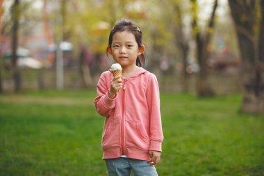 5-6 yaşlarında güzel bir Asyalı kız yaz günü parkta durup elinde lezzetli bir dondurma külahı tutuyor ve doğrudan kameraya bakıyor.. 