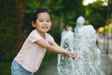 5-6 yaşlarında Asyalı bir kızın açık hava portresi. Günlük pembe tişört ve kot pantolon giyiyor. Yazın serin spreyin tadını çıkarırken çeşmede suyla gülüyor ve oynuyor.
