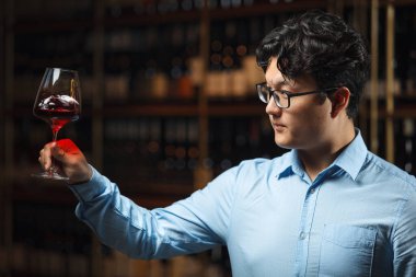 Profesyonel şarap garsonu, kırmızı şarabın berraklığını ve rengini inceliyor, bardağı ışığa odaklıyor ve çeşitli şarap şişeleriyle dolu arka plandaki mahzene dikkat ediyor..