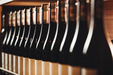 Şarap şişelerinin yakın plan görüntüsü, farklı marka ve çeşitleri sergiliyor, ahşap raflarda düzenli, yumuşak ışıklandırma ahşap kehribar tonlarını vurguluyor ve sıcak bir hava veriyor.