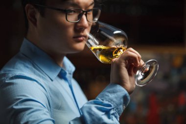 Odaklanmış Asyalı sommelier beyaz şarabın kokusunu dikkatlice değerlendiriyor, bardağı burnuna dayayıp, karanlık arka plandaki resmi kapatıyor..