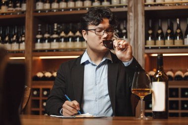 Asya kökenli yetenekli sommelier 'in kapalı mekan portresi kör şarap tadımı yapıyor. Yetenek seviyesini ölçüyor ve degulasyon listesine not alıyor. Mahzende dolu ahşaba karşı masada oturuyor.