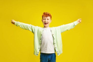 Studio portrait of happy little boy has a red hair cheerfully spreads arms to the sides, expressing joyful mood, wearing in stylish colorful clothes standing on yellow isolated background with a free clipart