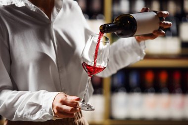 Kadın barmen tatmak için bardağa kırmızı şarap dolduruyor. Profesyonel kadın şarap garsonu, rafta duran şarap bardağına alkollü içecek doldurur..