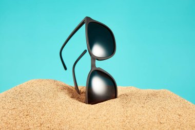 Güneş gözlükleri, siyah çerçeveli gölgeler, kısmen kumlara gömülü lensler mavi arka planda. Yaz mevsiminde, plajda, güneşli, turistik ve seyahatli. Yan görüş. Mesaj için yer. Boşluğu kopyala.