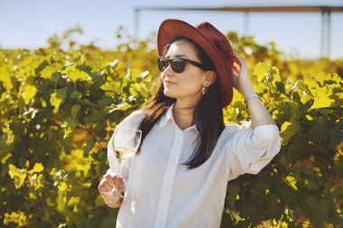 Sommelier kadını elinde şapka, elinde güneş gözlüğü, elinde şarap kadehiyle bir üzüm bağında duruyor..